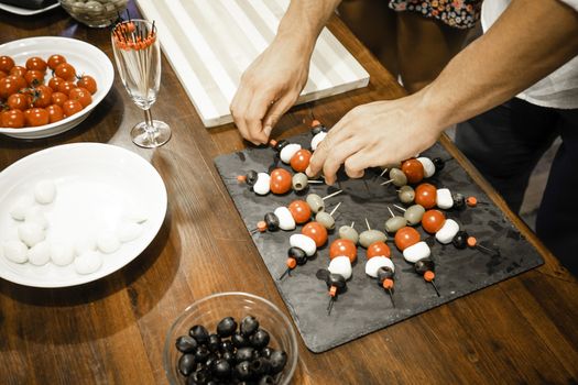 Top view of the arms of a young chef preparing vegetarian skewers appetizers in a slate plate on a wooden table with mozzarella, cherry tomatoes and olives - Happy hour at home with healthy food