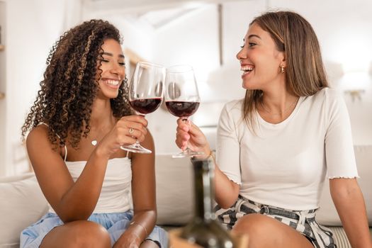 Two beautiful young women, Hispanic and Caucasian, sitting on a sofa toasting with a glass of red wine - Multiethnic happy female friends celebrating