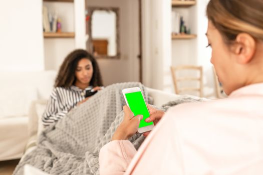 Two young mixed race female friends always connected communicating wearing pajama snuggle up on the sofa under a blanket - Selective focus on the green screen of the white smartphone