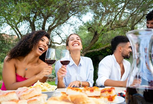 Mixed group of young happy friends celebrating outdoor with snacks and alcoholic drinks - Selective focus on the two cute smiling young hispanic and caucasian women toasting with red wine