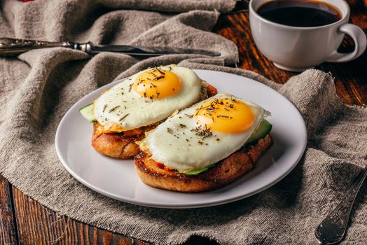 Toasts with vegetables and fried eggs on white plate and cup of coffee over grey rough cloth.