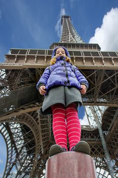A girl against the backdrop of the Eiffel Tower dressed in a blue jacket and red tights stands on a pillar. View from below.