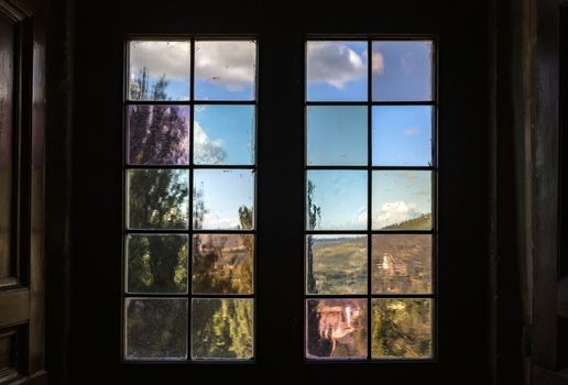 A window with multi-colored glass that overlooks a park or garden. Outside the window you can see a beautiful landscape of trees and the sky with clouds. Old wooden window with shutters
