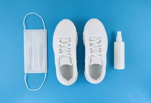 White medical mask, trainers, and hand sanitizer on blue background. Monochrome flat lay. Quarantine outfit.