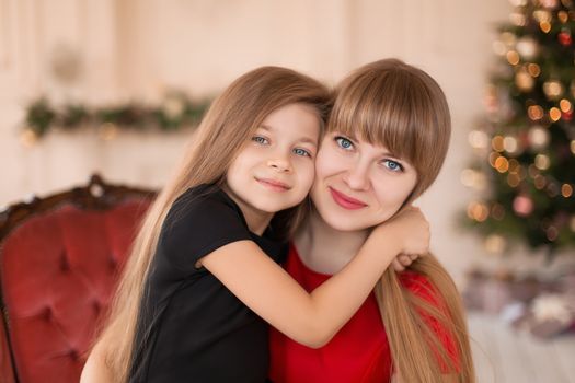 Little girl hugs her mom near the Christmas tree. Joyful moments of a happy childhood.