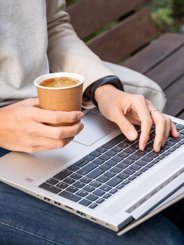 Business woman sits on wooden bench in urban park with laptop and cardboard cup of coffee. Student learns remotely from outdoors. Modern Internet technologies.