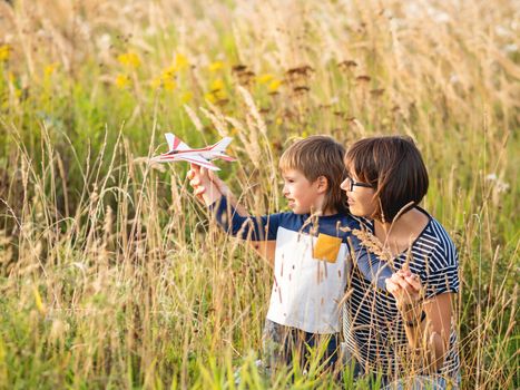 Cute boy and his mother play with toy air plane. Happy kid dreams to be a pilot. Boy is planning for the future. Mom and son on field at golden sunset hour at autumn season.