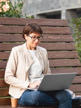 Business woman with short haircut and eyeglasses sits on wooden bench in park with laptop and cardboard cup of coffee. Student learns remotely from outdoors. Modern Internet technologies. Video call.