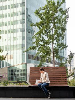 Business woman with short haircut and eyeglasses sits on bench in urban park with laptop and cardboard cup of coffee. Student learns remotely from outdoors. Modern Internet technologies. Video call.