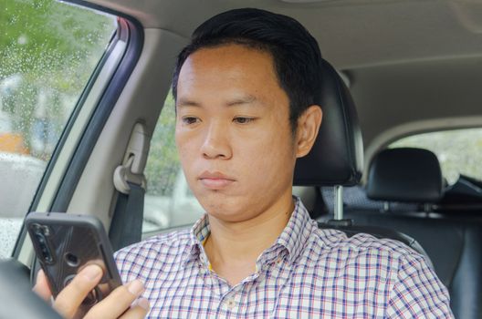 Asian man wearing a plaid shirt looks at smart phone in a car.