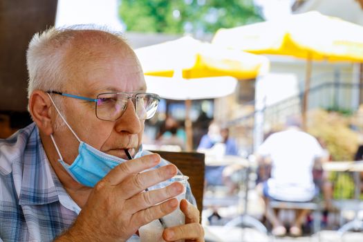 Old man in a medical face mask are a drinks water in summer cafe coronavirus covid-19