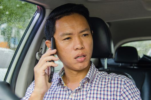 Asian man wearing a plaid shirt looks at smart phone in a car.