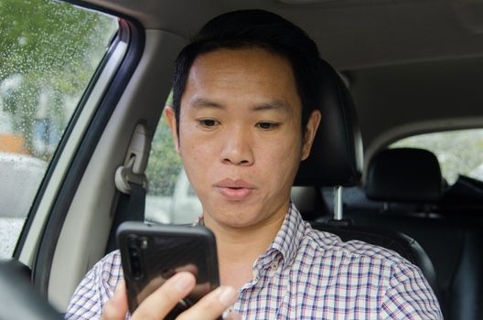 Asian man wearing a plaid shirt looks at smart phone in a car.