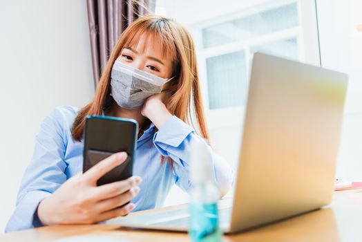 Asian young business woman wearing face mask protective using smart mobile phone for reading SMS message near laptop computer at home office quarantines disease coronavirus or COVID-19, work from home