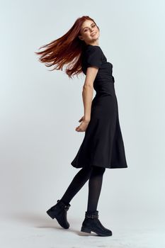 A woman in a black dress on a light background and pantyhose shoes red hair and pose in full growth. High quality photo