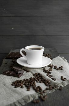 Coffee cup with cookies and chocolate with scattered coffee beans on linen and wooden table background. Mug of black coffee