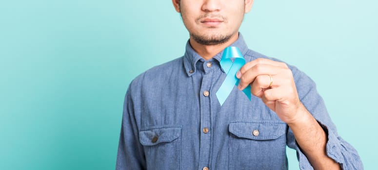 Asian portrait happy handsome man posing he holding light blue ribbon for supporting people living and illness, studio shot isolated on blue background, Prostate Cancer Awareness in November concept