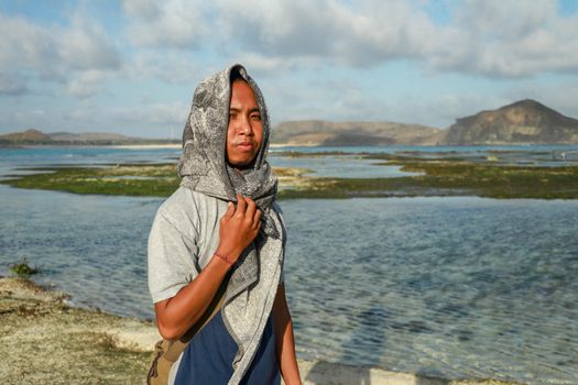 Close up teenage boy with towel on the head in casual clothes. A Guy on the beach. The guy in the towel at sea. Young asian man looks at the landscape.