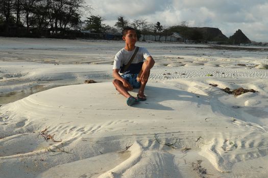 Man thinking and watching the sea on the beach. Teenager sitting on white sand at Tanjung Aan beach, Lombok, Indonesia. A young Asian man looks at the landscape.