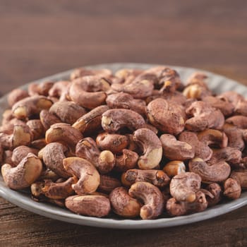 Cashew nuts with peel in a plate on wooden tray and table background, healthy raw food concept.