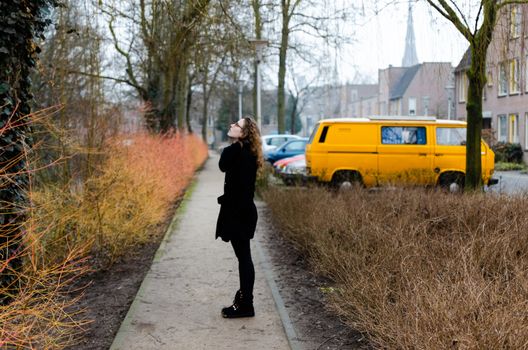 Photograph of a Spanish blonde girl dressed in black walking through the winter streets of a Dutch town with changing vegetation of yellow, orange, green and brown.
