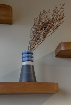 Dried flowers in beautiful ceramic blue vase standing on wooden shelf on wall of living room. Home decor. Space for text. Selective focus.