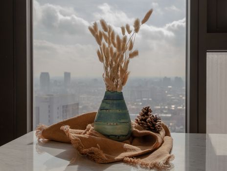 Dried flowers in beautiful ceramic blue vase handmade with two pine cones on marble table of living room and city view. Home decor. Space for text. Selective focus.
