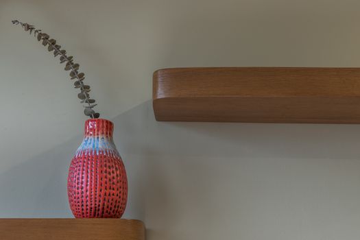 Dried flowers in beautiful ceramic red vase standing on wooden shelf on wall of living room. Home decor. Space for text. Selective focus.