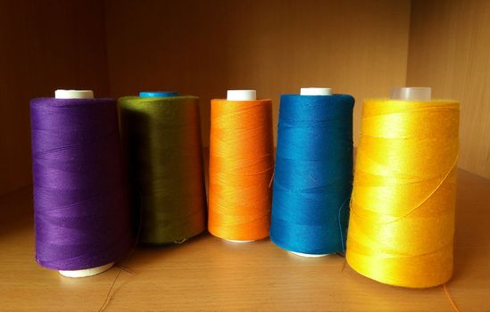 Multicolored sewing threads in bobbins stand on a brown shelf.