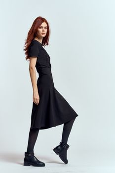 A woman in a black dress on a light background and pantyhose shoes red hair and pose in full growth. High quality photo