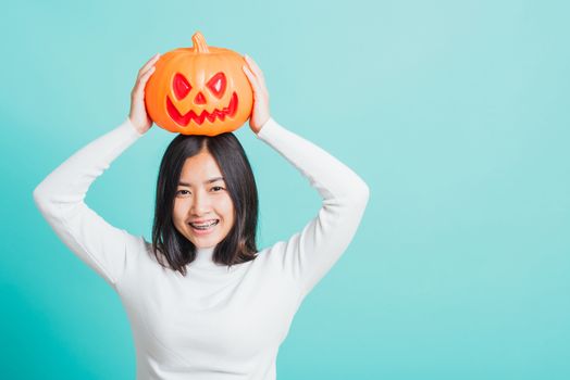 Portrait of Asian beautiful young woman holding orange model pumpkins and put it on the head, funny happy female with ghost pumpkins, studio shot isolated on blue background