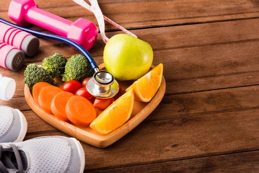 Top view of fresh fruits and vegetables in heart plate wood (apple, carrot, tomato, orange, broccoli) and sports equipment and doctor stethoscope on wooden table, Healthy lifestyle diet food concept