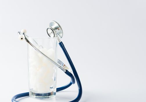 A glass full of white sugar cube sweet food ingredient and doctor stethoscope, studio shot isolated white background, health high blood risk of diabetes and calorie intake concept and unhealthy drink