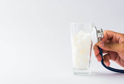 Hand of doctor hold stethoscope check on glass full of white sugar cube sweet food ingredient, isolated on white background, health high blood risk of diabetes and calorie intake unhealthy drink