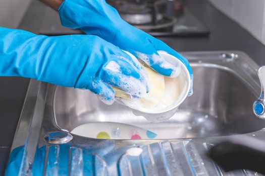 Close up of a man wearing a blue rubber glove was washing the cup with a dish washing liquid in the sink in the kitchen.