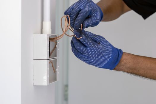 The electrician wears blue gloves, is using an electric cable cutter knife to install the plug and power switch on the wall.