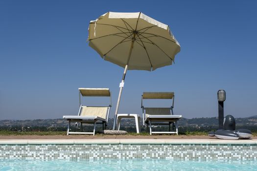 Blue swimming pool with beach lounger on wooden flooring with parasol, sun deck on sea view for summer vacation