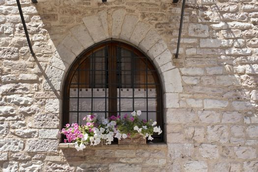 A basket of bright pink and red flowers hangs on the window of a home in an ancient building in Italy, surrounded by beautiful patterns in the stone wall.