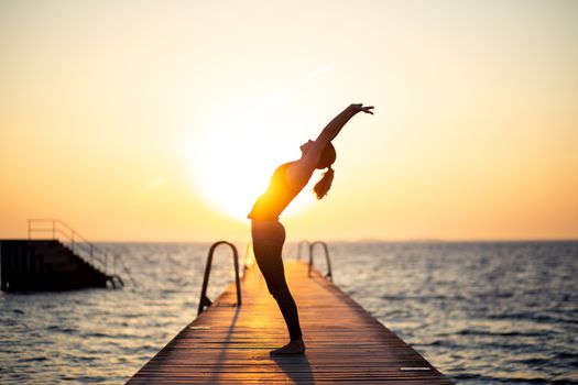 Silhouette of a woman practizing yoga on a wooden pier during sunset.