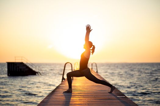 Silhouette of a woman practizing yoga on a wooden pier during sunset.