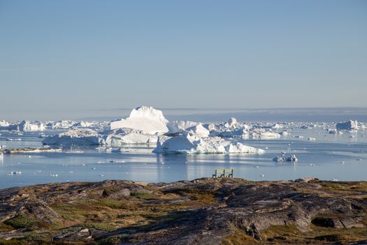 Ilulissat, Greenland - July 9, 2018: A bench at the coastline with iceberg view. Rodebay, also known as Oqaatsut is a fishing settlement north of Ilulissat.