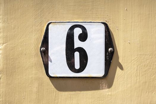 Number 6 in white on a black metal plate on a yellow concrete wall