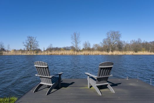 Rustic hand crafted chairs on a wooden deck overlooking a river or lake with reeds in spring sunshine