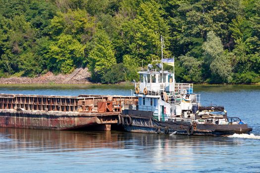 River tug slowly pushing an empty rusty barge down the river against coastal greenery, river freight transport concept, copy space.