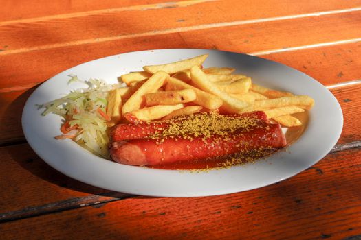 Traditional German currywurst, served with chips on a white plate. Orange wooden table as background.
