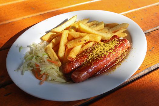 Traditional German currywurst, served with chips on a white plate. Orange wooden table as background.