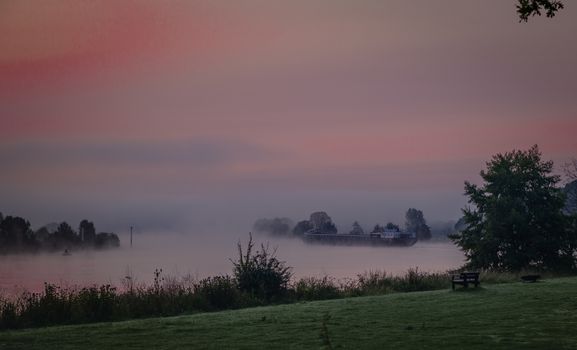 ship in the maas during sunrise in the early morning in limburg in holland with the trees mist and hazy fog