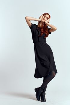 A woman in a black dress on a light background and pantyhose shoes red hair and pose in full growth. High quality photo