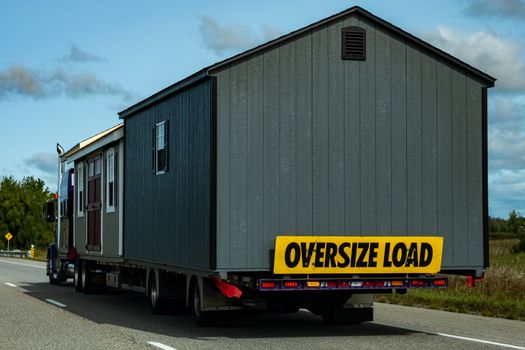 Two pre-built garden sheds are being transported by a semi-trailer transport truck on a highway, with a sign on the back indicating an oversize load.