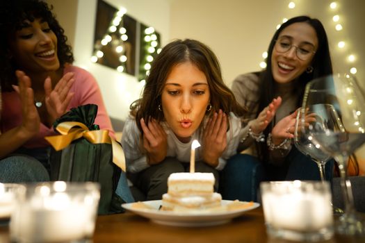 Close up of cute young woman blows on the cake candle next to her present celebrating her birthday at home with her two mixed race hispanic and caucasian female best friends